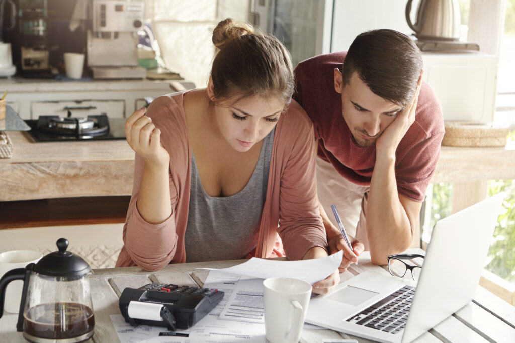 Young married couple with many debts doing paperwork together, reviewing their bills, planning family budget and calculating finances at kitchen table with papers, calculator and laptop computer